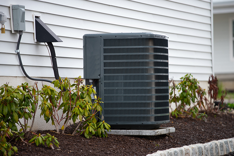 Air conditioner beside a home with green bushes.