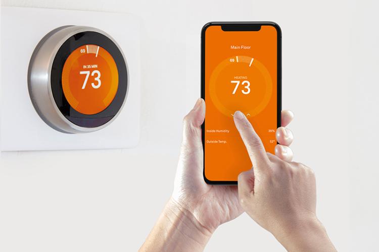 Hand holding up a smartphone next to a thermostat and controlling the temperature from the phone.